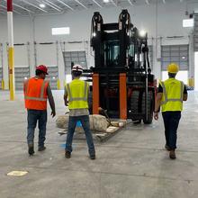 Three people in hard hats and other safety gear stand in front of a forklift carrying a piece of concrete inside a warehouse.