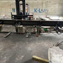 Inside a warehouse, a steel beam is attached to a concrete column being lifted by a forklift.