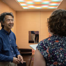 Yoshi Ohno speaks to Megan King in a small room with a specialized ceiling light fixture showing different colors of light. 