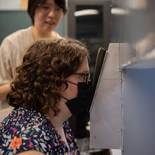 Megan King wears a black mask as she looks into a lighting test device, with Jane Li standing in the background. 