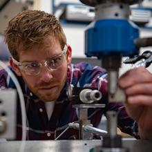 Jake Benzing wears safety glasses as he bends over to adjust piece of scientific equipment in the lab.