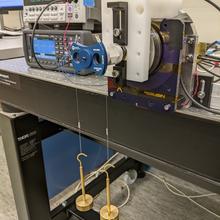 Scientific equipment on a lab table includes stacked boxes with readouts and device with two weights suspended by wires.