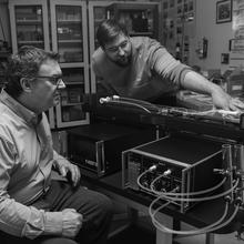 Black and white "recreation" photo shows Jim Norris seated next to a scientific instrument while Peter Trask leans over a horizontal component. 