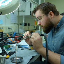 Peter Trask sits at a lab table working on wiring for a scientific device. 