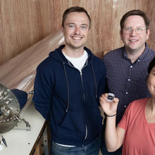 A smiling woman stands with two smiling men, holding up a small device. Two their right is a large cylindrical metal instrument, lying horizontally.