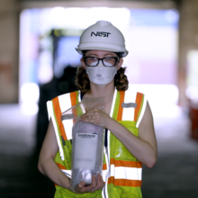 A woman in a NIST hard hat and other safety gear stands inside a warehouse holding a plastic bag labeled "Evidence."