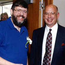 Two men standing side by side smiling, the one on the left with a single white flower pinned to his polo shirt, the one on the right in a suit and tie.