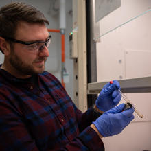 A man wearing safety goggles and rubber gloves inspects a test tube he is holding alongside a magnet. 