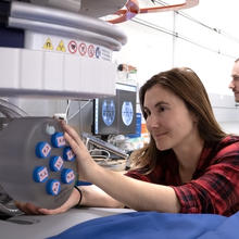 A woman leans over an MRI machine to reach a set of samples in a round holder, while a man sits behind her at a computer looking at MRI images. 