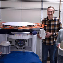 Three casually dressed researchers stand smiling around an MRI machine, each holding a different device used in their experiments.