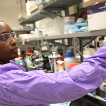 Alshae' Logan-Jackson wears a purple coverall and safety glasses in the lab as she uses tweezers to hold a small white disk. 