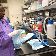Alshae' Logan-Jackson wears a purple coverall and safety glasses as she stands at a lab table holding a plastic sample bag. 