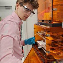 Win Lawson wears safety glasses as he stands at a lab table with an open cabinet made of translucent orange-tinted plastic.