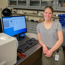 Grace Waters poses smiling in the lab, leaning against a desk with a computer monitor showing data and graphs. 