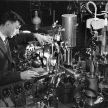 A black and white image of a man in a gray suit standing next to a complex machine with a profusion of tubes, wires and lights.