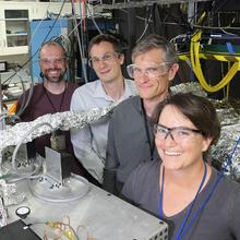 Four researchers pose standing in a lab, smiling and wearing safety glasses, surrounded by wires and other electronics.