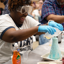 Kandice Taylor, in safety glasses, laughs as she pours liquids into a glass flask that is overflowing with foam.