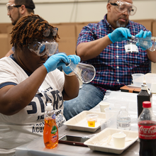 Teachers at a seminar wear safety glasses and look into glass flasks while sitting at a table of experiment supplies. 