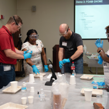 Workshop participants stand around a table donning safety gear like glasses and gloves as they prepare to practice an experiment.