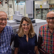 Three researchers pose smiling in the lab, standing in front of a large piece of equipment.  