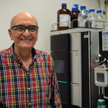 Miral Dizdaroglu wears safety glasses as he poses smiling in front of a large piece of lab equipment.