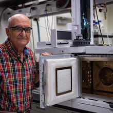Miral Dizdaroglu wears safety glasses as he poses in the lab next to an open spectrometer, which looks a little like a microwave with extra buttons. 