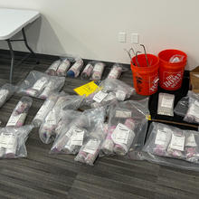 Packets of cylindrical objects wrapped in clear plastic bags with white labels are lined up on the floor of a conference room.