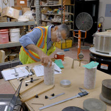 A researcher wearing safety goggles and a high-viz vest leans over to examine a concrete core sample on a lab table.