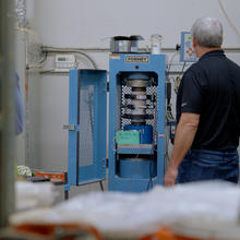 A researcher faces away from the camera toward a concrete core sample held vertically in a blue metal testing machine. 