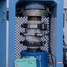 A concrete core sample shatters as it is held vertically inside a blue metal testing machine. 