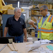 Two men stand a table in a warehouse space, looking at a broken piece of concrete that one is holding. 