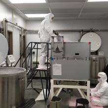A researcher in a white coverall stands on a stool feeding material into a large square machine putting off cold vapor, while another researcher sits at the bottom to collect material coming out. 