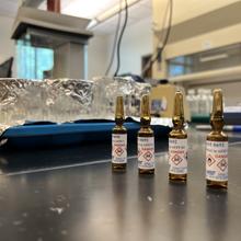 Four small dark glass ampules stand on a lab table, marked with skull and crossbones danger icons. 