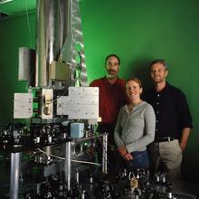 Two men and a woman pose standing behind a large scientific device with a metal cylinder reaching to the ceiling and green light shining out. 