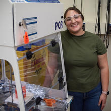 Woman in safety glasses stands next to an enclosed space with scientific equipment inside.