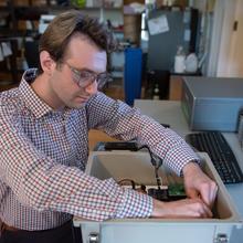 A man wearing safety glasses and sitting at a computer workstation reaches into a box with wiring and circuitry.