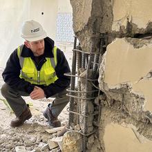 A man in safety gear crouches to examine a damaged wall inside a building after an earthquake. 