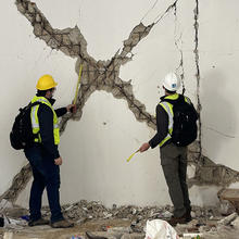 Two people wearing safety gear and backpacks and holding tape measures face away from the camera toward a wall with an X-shaped damaged area. 