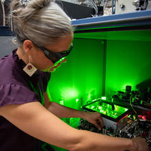 A researcher wearing safety glasses reaches into a box of circuitry and other equipment, which emits a green glow. 