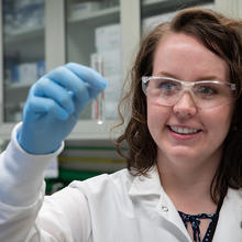 Jerilyn Izac, wearing safety glasses and a lab coat, holds up test tube containing clear liquid.