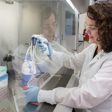 Jerilyn Izac sits at a lab bench with her hands inside a plastic shield, using a device to draw liquid from a plastic container into a tube. 