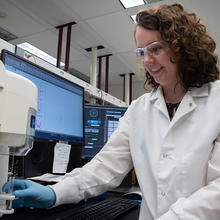 Jerilyn Izac, wearing safety glasses and a lab coat, places a test tube into a holder on a large piece of lab equipment.