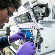 A researcher leans close to an open boxlike device, handling small tubes and vials inside.