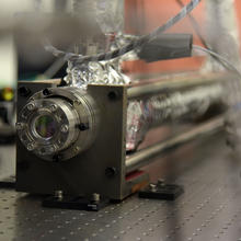 A scientific device attached to a lab table has a round, porthole-like opening on one end of a cylinder wrapped in foil. 