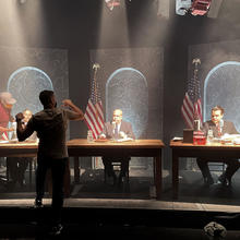A man faces away from the camera, gesturing as he speaks to three actors seated behind desks on a film set. 