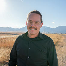 Zach Grey poses outdoors with wind turbines in the background. 