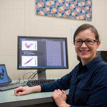 Melinda Kleczynski poses sitting at her desk, with a monitor showing data and graphs behind her. 