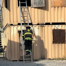 A firefighter climbs a ladder into a shipping container, while a person to their left tracks their location.