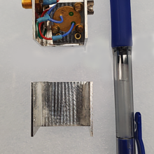 A square device with wiring inside is taken apart for viewing with a pen laid alongside for scale. 