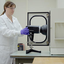 Amy Mensch, wearing a lab coat and gloves, adjusts a black camera-like device in the lab. 